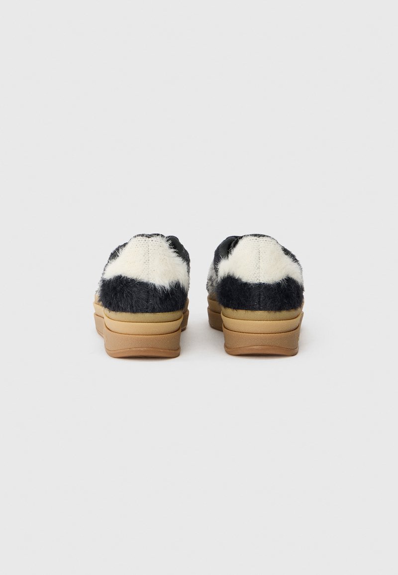 Hausschuhe aus Kunstfell mit einem schwarz-weißen Kuhmuster, brauner Gummisohle und gepolstertem Absatz. Kompaktes Design mit runder Zehenpartie.