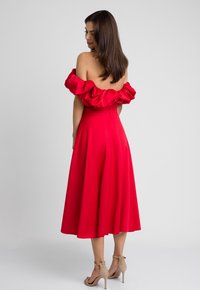 Czerwona sukienka na jedno ramię z falbaniastym dekoltem, zwiewnym dołem w kształcie litery A i gładką teksturą. Noszona z jasnobeżowymi szpilkami.