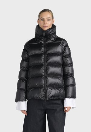 Femme portant une doudoune noire brillante avec un col haut, des poignets de chemise blancs visibles et un pantalon foncé, debout devant un fond uni.