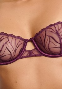 Soutien-gorge violet avec mesh transparent orné de broderies en forme de feuilles, armatures, bords festonnés et petite découpe centrale.