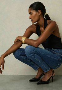 Bodycon nero con scollo a halter, jeans di denim blu, bracciale a manica d'oro, décolleté neri a punta. Modella accovacciata contro uno sfondo neutro.