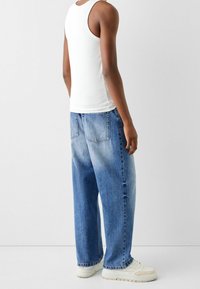 Vit linne, ljusblå löst passande denimjeans med blekt utseende, bakfickor synliga, bärs med beige sneakers. Neutral bakgrund.