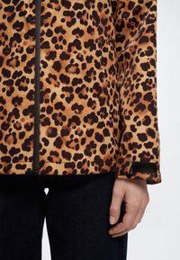 Leopárdmintás dzseki fekete cipzárral és tépőzáras mandzsettával, fehér háttér előtt, kézzel látható személy által viselve.