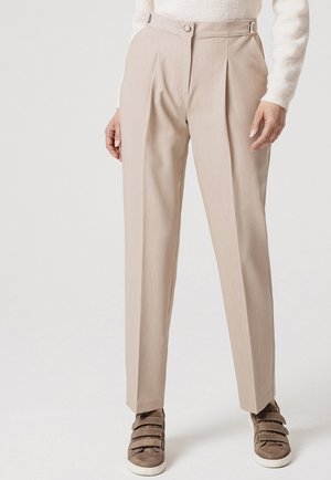 Pantaloni beige a vita alta su misura con pieghe, indossati con un maglione crema e sneakers marroni con chiusura in velcro.