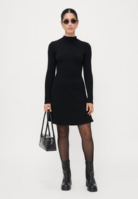 Robe noire en tricot côtelé avec un col montant, des manches longues, un ourlet évasé, assortie avec des bottes mi-mollet et un petit sac à main.