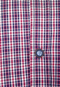 Rood, wit en blauw geblokt stof met een knoopdetail aan de voorkant van een shirt, met dunne verticale en horizontale lijnen in een ruitpatroon.
