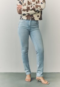 Svijetloplave uske traperice s glatkom teksturom, uparene s smeđim i kremastim džemperom s uzorkom i rebrastim rubom. Sandale s tankom potpeticom.