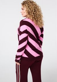 Vrouw met krullend haar, gekleed in een roze en donkerpaarse gestreepte trui en donkerpaarse broek met zijstrepen, zijwaarts gericht tegen een witte achtergrond.