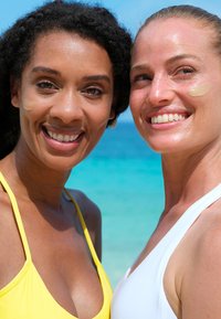 Due donne sorridenti in spiaggia. Una indossa un costume da bagno giallo, l'altra uno bianco. Entrambe hanno una leggera crema applicata sul viso.