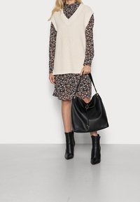 Krémová pletená vesta s výstřihem do V přes černé šaty s květinovým vzorem. Celý vzhled doplňuje černá kožená kabelka a černé kotníkové boty.