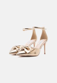 Χρυσά μεταλλικά ψηλοτάκουνα παπούτσια με μυτερές μύτες, λεπτομέρεια κορδέλας στο μπροστινό μέρος και λουρί αστραγάλου με αγκράφα. Λεία υφή.
