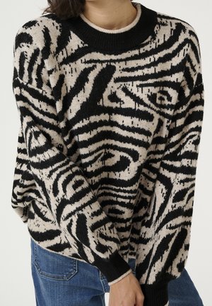 Person trägt einen schwarz-beigen Pullover mit abstraktem Muster, kombiniert mit blauen Jeans, steht vor einem einfarbigen hellen Hintergrund.