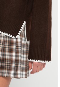Καφέ πουλόβερ με ριμπ υφή και λευκή περίμετρο με κυματιστό σχέδιο, σε συνδυασμό με μια καρό φούστα σε καφέ, λευκές και μαύρες αποχρώσεις.