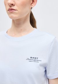 Γυναίκα που φοράει ανοιχτό μπλε t-shirt GANT με λογότυπο στο αριστερό στήθος, τα μαλλιά της είναι πιασμένα πίσω και φοράει μικρό κρίκο στο αριστερό αυτί.