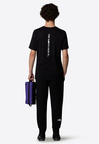 Sort bomulds T-shirt med hvid lodret branding på ryggen, parret med sorte sweatpants og en lilla justerbar taske.