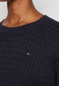 Marineblauer gestrickter Pullover mit Kabelmuster, Rundhalsausschnitt und einem kleinen roten, weißen und blauen Logodetail auf der Brust.
