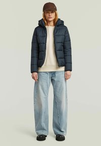 Dark blue puffer jacket with a zip closure and hood, worn over a light beige sweater and loose light denim jeans, complemented by a brown cap.