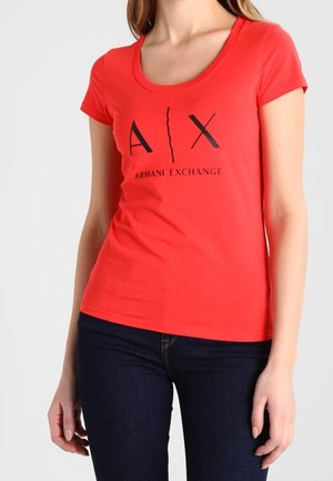 Kobieta ubrana w dopasowany czerwony t-shirt Armani Exchange z czarnym logo, w połączeniu z ciemnoniebieskimi dżinsami, stojąca na jednolitym tle.