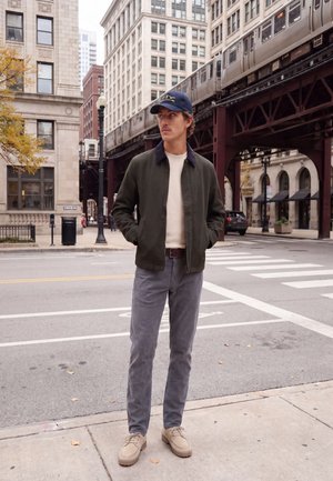 Un homme portant une casquette bleu marine, une veste sombre, un pantalon gris et des chaussures beiges se tient sous une voie ferrée aérienne, dans une rue de la ville.