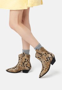 Botines de piel de serpiente marrón con punta afilada y tacón bajo. Presentan un patrón texturizado y calcetines grises, posicionados sobre un fondo blanco.
