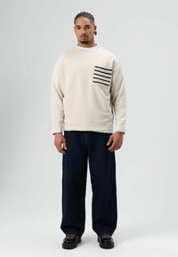 Beige oversized sweater met een gestreept zakje, gecombineerd met donkerblauwe wijde jeans en zwarte schoenen met dikke zolen.