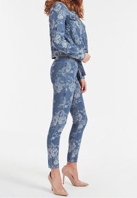 Giacca in denim e jeans coordinati in blu con motivi floreali bianchi. La giacca ha maniche lunghe e due tasche sul petto. Jeans slim-fit.