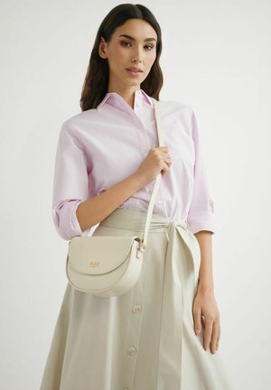 Kobieta ubrana w jasnoróżową koszulę w paski i beżową spódnicę na guziki, trzymająca małą beżową torebkę na ramię z klapką i złotym logo.