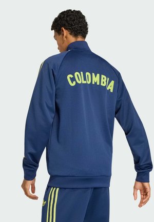 Mann trägt marineblaue Sportjacke und Hose mit gelben Streifen, "COLOMBIA" auf der Rückseite der Jacke gedruckt.