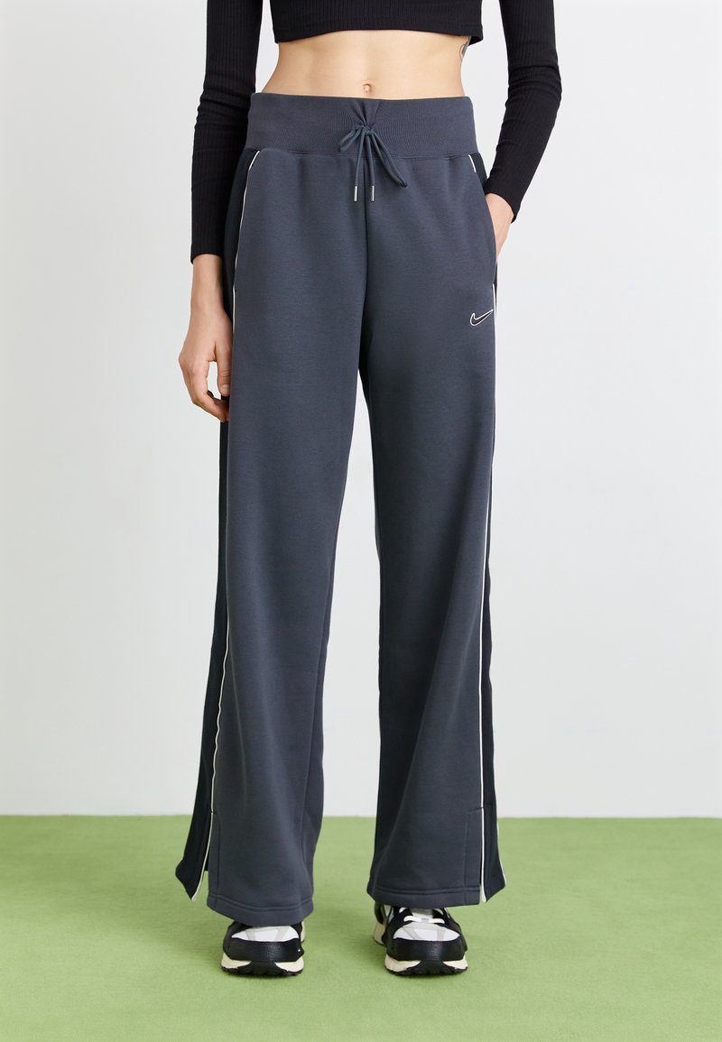 Osoba oblečená v tmavě šedých teplácích Nike s bílými bočními pruhy, černobílých teniskách a černém zkráceném dlouhém rukávu na zelené podlaze.