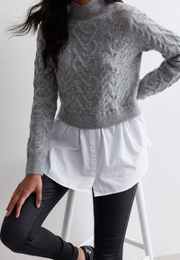 Grauer kabelgestrickter Pullover mit hohem Kragen, kombiniert mit einem weißen Hemd mit Knöpfen und schwarzen Leggings, sitzt auf einem Hocker.