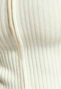 Kremowy prążkowany materiał z pionowymi paskami, wyposażony w ukryty zamek błyskawiczny po jednej stronie. Miękka tekstura i równomierne przeszycia.