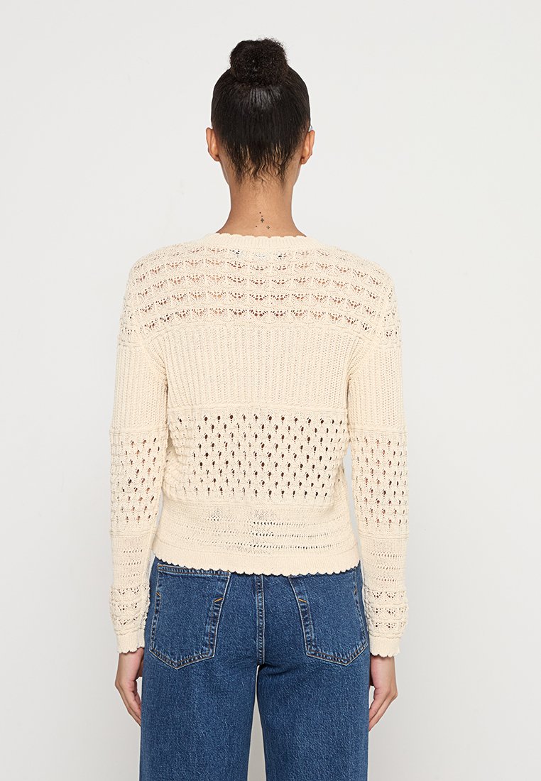 Jasnobeżowy sweter dzianinowy z teksturowanym wzorem ażurowym, prążkowanym wykończeniem i długimi rękawami. Połączony z niebieskimi dżinsami z wysokim stanem.