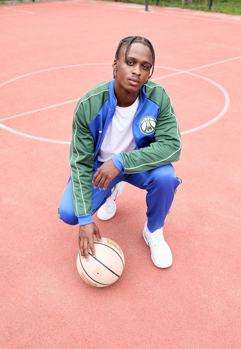 Blue and green tracksuit with basketball logo, white T-shirt, and white sneakers. Individual crouching on a basketball court holding a ball.