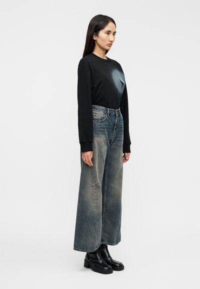 Cămașă neagră cu mâneci lungi și design în degrade, combinată cu blugi largi din denim de culoare albastru estompat, cu accente uzate, purtați cu cizme negre.