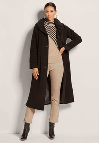 Zwarte lange jas met een brede kraag, knoopsluitingen, gedragen over een gestreepte beige en zwarte top en tan broek, gecombineerd met zwarte enkellaarsjes.
