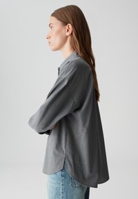 Gray button-up shirt with a relaxed fit, featuring a slightly curved hem, long sleeves, and a textured fabric. Worn over light blue jeans.