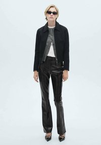 Zwarte cropped jack over een grijs overhemd met knopen en een witte top, gecombineerd met glanzende zwarte broeken en puntige hakken. Zonnebril maakt de look compleet.