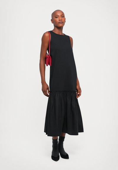 Schwarzes ärmelloses Kleid mit gerafftem unteren Saum, aus glattem Stoff gefertigt. Kombiniert mit einer roten Handtasche und schwarzen Socken mit spitzen Absätzen.