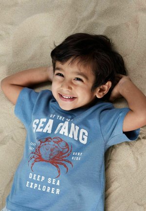 T-shirt en coton bleu avec un graphique de crabe rouge et un texte blanc indiquant "EXPLORATEUR DES PROFONDEURS" et "GANG DES MERS", porté par un enfant allongé sur le sable.