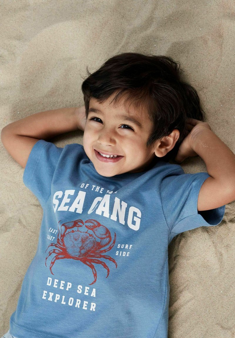 Blå bomulds t-shirt med et rødt krabbe-design og hvid tekst, der læser "DEEP SEA EXPLORER" og "SEA GANG", båret af et barn, der ligger på sandet.