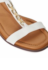 Witte textuur sandalen met een platte zool en een bruine binnenzool, voorzien van gouden kettingaccenten op de T-sandalen. De banden zijn slank en minimalistisch.