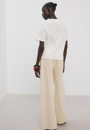 Femme avec un chignon portant un haut blanc à manches courtes, un pantalon beige à jambes larges et plusieurs bracelets, debout devant un fond clair uni.