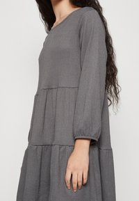 Grå, texturerad långärmad klänning med ett lager-på-lager-design och rundad halsringning. Har voluminösa ärmar och två sidofickor.