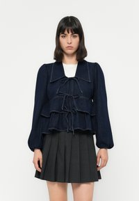Denim peplum top in een donkerblauwe kleur, met gegolfde lagen, een brede kraag en een gestrikte sluiting aan de voorkant. Licht geprofileerde stof met pofmouwen.