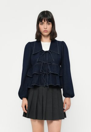 Denim peplum topp med en mørk blå farge, volanger, bred krage og knytefront. Lett teksturert stoff med puffede ermer.