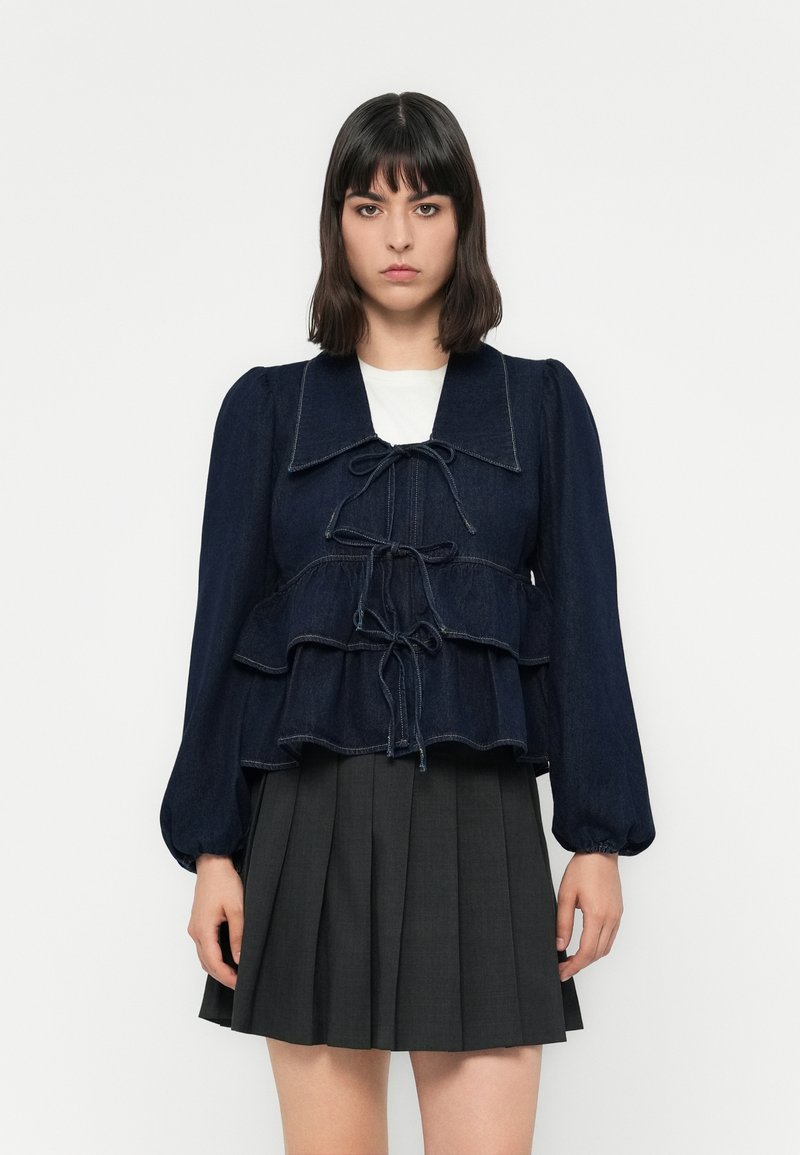 Denim peplum top with a dark blue color, ruffled layers, a wide collar, and tied front closure. Lightly textured fabric with puffed sleeves.