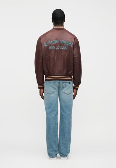 Καφέ δερμάτινο μπουφάν bomber με ριμπ μανσέτες και τελείωμα, με κεντημένο μπλε λογότυπο "Emporio Armani Eagle Club" στην πλάτη. Φορεμένο με ανοιχτόχρωμο μπλε τζιν.