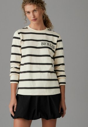 Chemise à rayures à manches longues en crème et noir avec "BERLIN" imprimé sur la poitrine ; associée à une jupe noire volumineuse.
