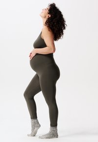 Gravid kvinde iført olivenfarvet tætsiddende tøj og grå sokker, stående på tå, holder om sin mave, ser opad med krøllet hår.