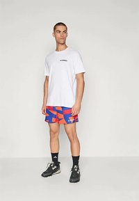 Baltas īsās piedurknes T-krekls ar melnu "TERREX" logo, savienots ar spilgti ziliem un sarkaniem rakstainiem šortiem. Melni sporta apavi un potīšu zeķes noslēdz izskatu.